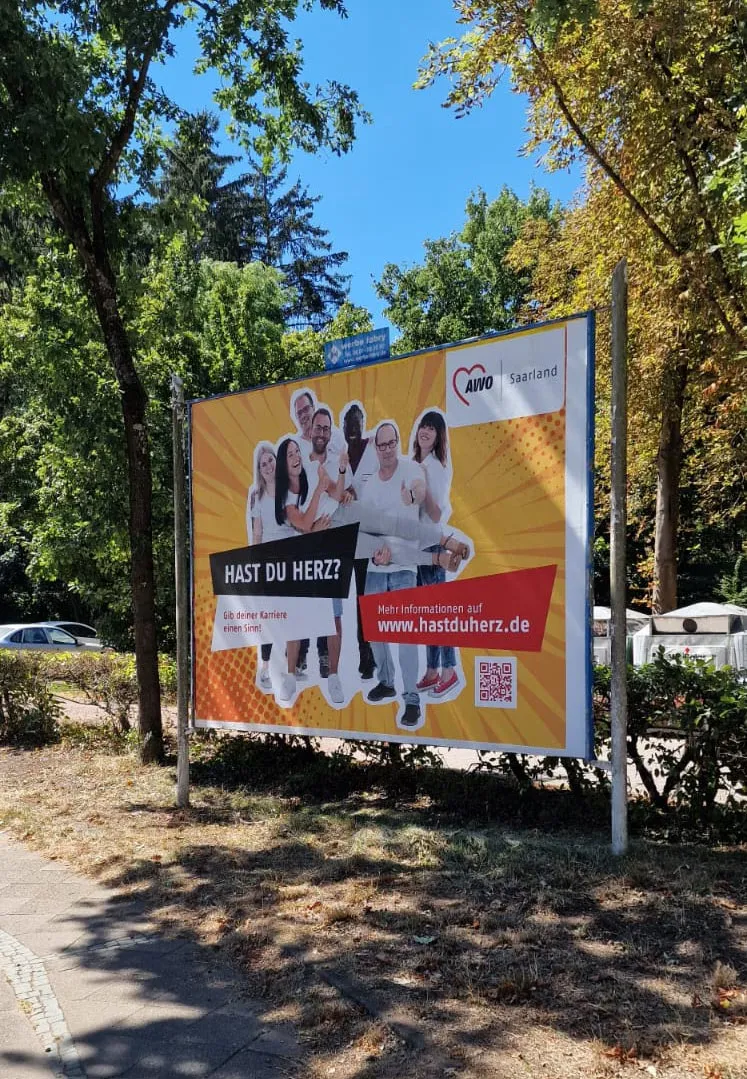 Großflächenplakat der AWO-Recruitingkampagne „Hast du Herz?“ am Straßenrand unter Bäumen