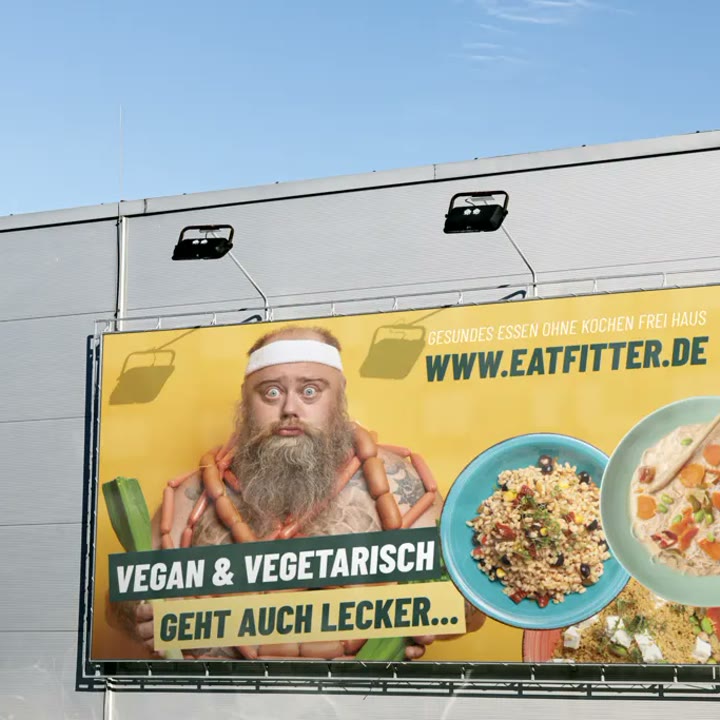 EATFITTER-Plakat an einer Hallenwand: bärtiger Mann mit Gemüse und Fertiggerichten, Slogan „Vegan & vegetarisch“