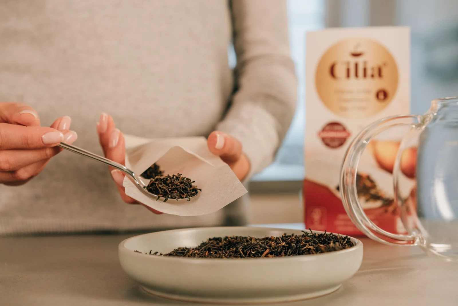 Produktfotografie für Cilia: loser Tee wird mit dem Löffel in einen Filter gefüllt, Teeverpackung im Hintergrund