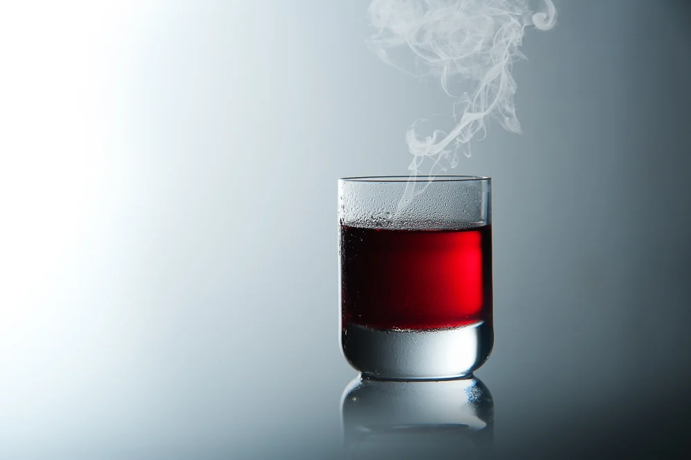 Produktfotografie: Glas mit dunkelrotem Getränk und aufsteigendem Rauch vor hellem Hintergrund