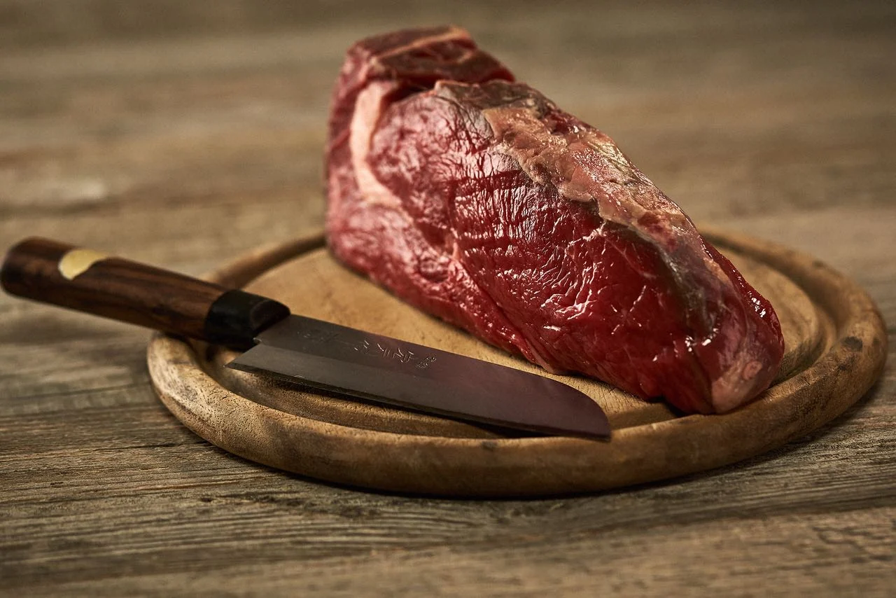 Foodfotografie für einen Fleischkatalog: Stück rohes Rindfleisch mit Messer auf rundem Holzbrett
