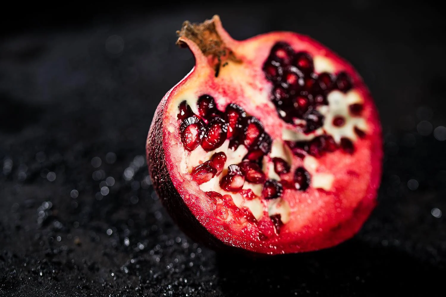Foodfotografie: aufgebrochener Granatapfel mit roten Kernen auf dunklem, nassem Untergrund