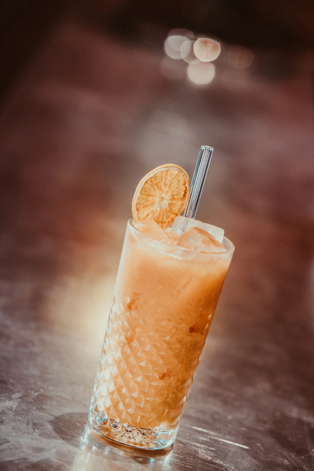 Getränkefotografie: orangefarbener Cocktail mit getrockneter Orangenscheibe und Strohhalm an der Bar