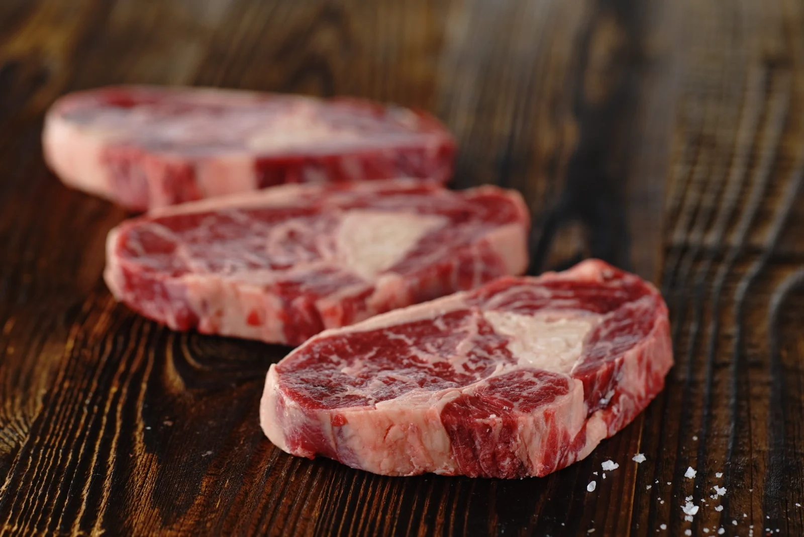 Foodfotografie: drei rohe Rib-Eye-Steaks mit Salz auf dunklem Holz
